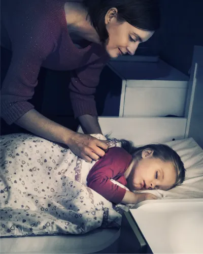 A mother tucks her young daughter into bed.
